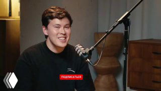 Улыбающийся участник подкаста в студии, говорит в микрофон во время записи интервью; видны микрофон, стойка и фон студии.