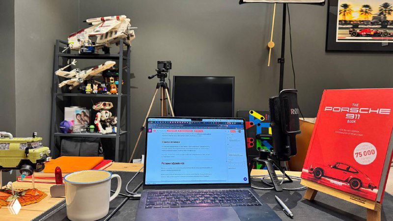 Рабочее место фрилансера: ноутбук с открытой страницей, камера на штативе, книжка про Porsche, конструктор и кружка на рабочем столе, предметы автора