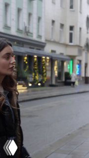 Превью вертикального ролика: молодая женщина в тёмной куртке идёт по городской улице с витринами — кадр для сравнения Reels.