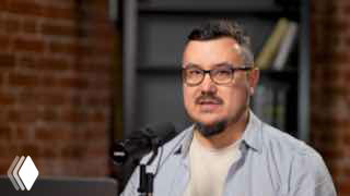 Фото ведущего в студии за микрофоном: мужчина в очках говорит в подкаст на фоне кирпичной стены и полок с книгами; крупный план, разговорная обстановка.