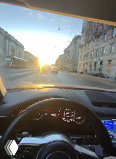 Вид с места водителя: рулевое колесо и панель приборов на фоне городской улицы и заходящего солнца, кадр POV за рулём.