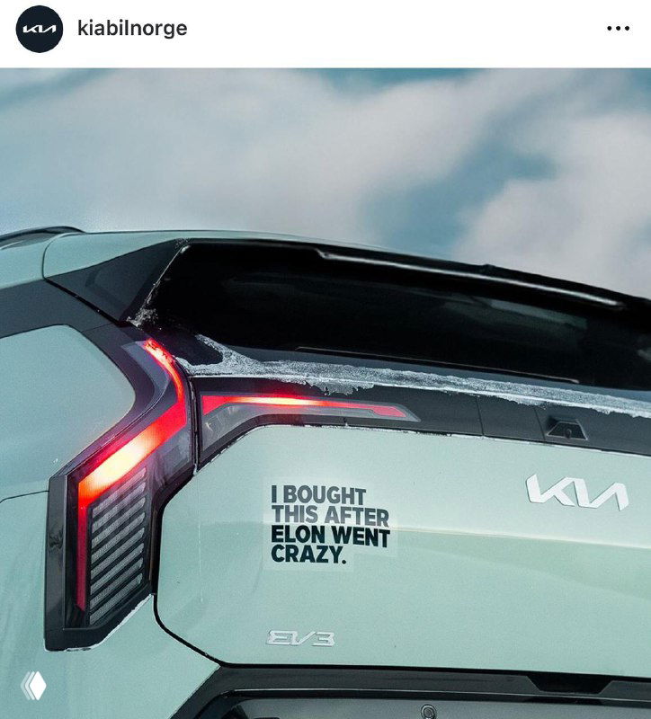 Задняя часть электрического KIA вблизи с наклейкой «I BOUGHT THIS AFTER ELON WENT CRAZY», виден логотип KIA и небо на фоне