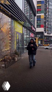 Человек с телефоном идёт по тротуару мимо витрины массажного салона на фоне жилых домов — вертикальная уличная сцена.