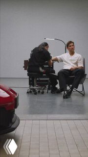 Мужчина сидит в кресле, ему делают татуировку в светлом помещении; на переднем плане видна часть красного автомобиля.