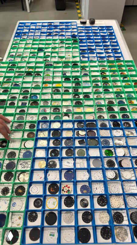 Вид сверху на стол с множеством циферблатов завода «Луч»: десятки лотков зелёного и синего цвета, каждый циферблат с уникальным принтом и оформлением.