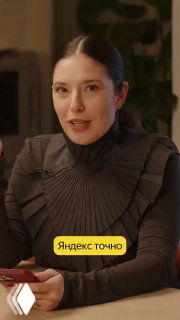 Кадр из видео: женщина в тёмной одежде сидит за столом в офисном интерьере, видно плакат/плашку с подписью «Яндекс точно», интервью в студии.