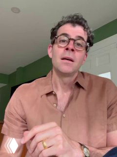 Портретный кадр вертикального видео: мужчина в очках разговаривает в комнате с зелёной стеной, выглядит как скриншот демонстрации функции перевода в соцсети.