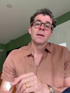 Вертикальный видеокадр: человек в коричневой рубашке и очках говорит в камеру на фоне зелёного интерьера — похоже на отрывок демонстрации перевода в приложении.