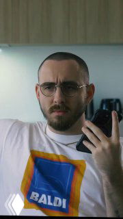 Портретный кадр мужчины в очках и футболке с ярким принтом — герой ролика демонстрирует эмоции при общении с клиентом в офисной обстановке.