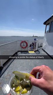 На палубе лодки рука держит банку Grillo's Pickles, вдалеке видна Статуя Свободы и городской горизонт — уличная дегустация огурцов.