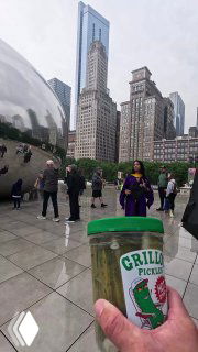 Рука держит банку Grillo's Pickles на фоне популярной городской достопримечательности и небоскрёбов; кадр подчёркивает уличный промо-формат бренда.