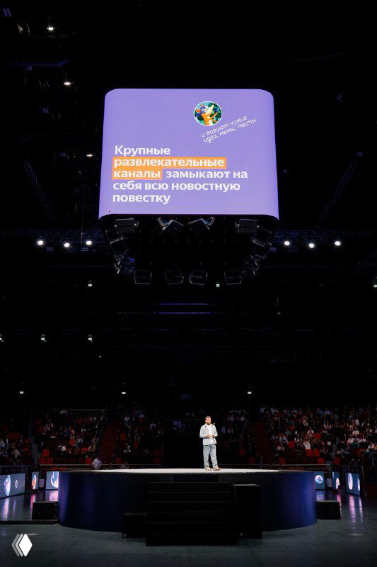 Докладчик на сцене конференции Яндекса перед большим экраном с слайдом о развлекательных каналах; затемнённый зал, сцена и аудитория.
