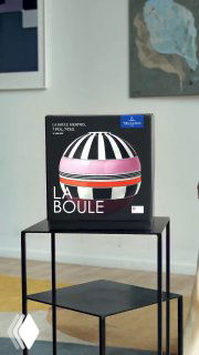 Фотография коробки с ярким дизайном La Boule, стоящей на металлической полке в интерьере с картинами на стене и частью помещения; кадр демонстрирует упаковку продукта на фоне комнаты.