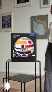 Кадр упаковки La Boule на той же металлической полке, вид с другого ракурса, в кадре видна часть человека и настенные картины; фотография иллюстрирует объект визуального внимания в ленте.