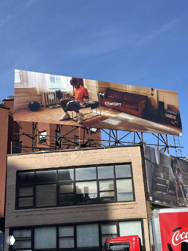 Фотография большого билборда над зданием с рекламой ChatGPT: сцена в гостиной, лёгкий ретро-стиль и логотип в правой части композиции