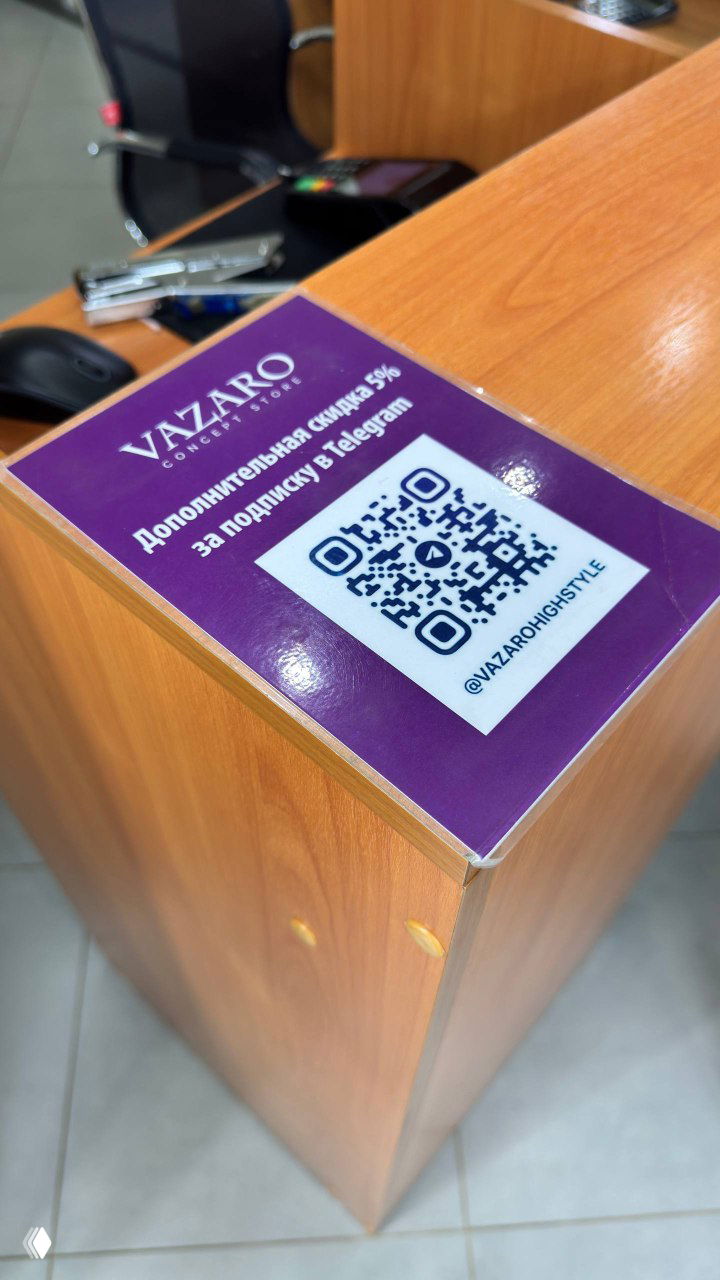 Фиолетовая наклейка на прилавке магазина с QR-кодом и надписью о дополнительной скидке 5% за подписку в Telegram; видна деревянная стойка кассы и логотип магазина Vazaro.