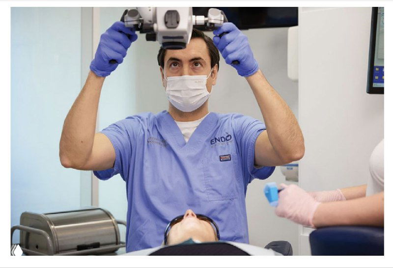 Врач в маске подводит операционный микроскоп к рабочему полю над пациентом, ассистент готовит инструменты рядом — демонстрация движения класса IV.