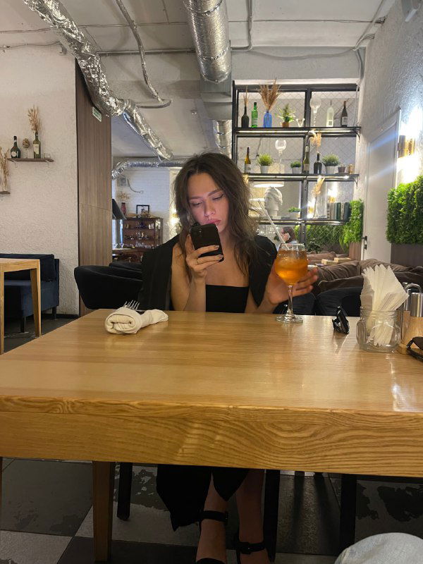 Женщина в кафе сидит за деревянным столом и смотрит в телефон; интерьер и поза используются как референс для переноса сцены и добавления объектов.