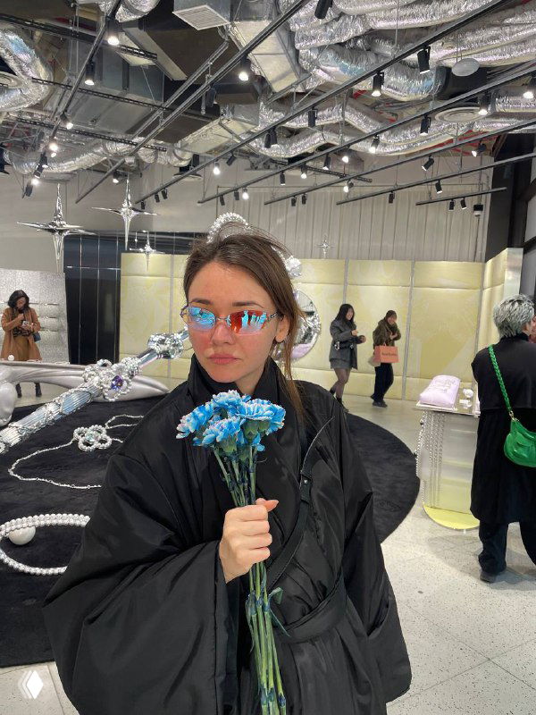 Женщина в тёмной одежде держит букет голубых хризантем на фоне выставки: инсталляции и прохожие видны в зале.