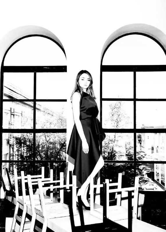 Чёрно-белая фотография женщины в платье, стоящей на стульях у больших арочных окон — образ решимости и лидерства.