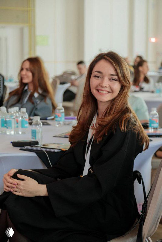 Улыбающаяся женщина в деловом наряде на конференции, за столами участники, блокноты и бутылки воды, атмосфера обучения и нетворкинга.