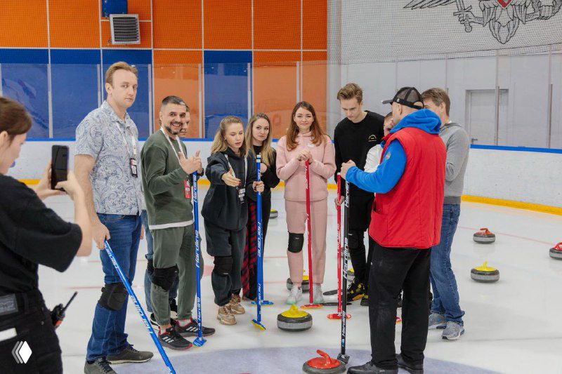 Группа людей на льду в спортивном зале во время тренировки по керлингу: участники с метлами и камнями позируют в зале, показывает командную работу и взаимодействие.