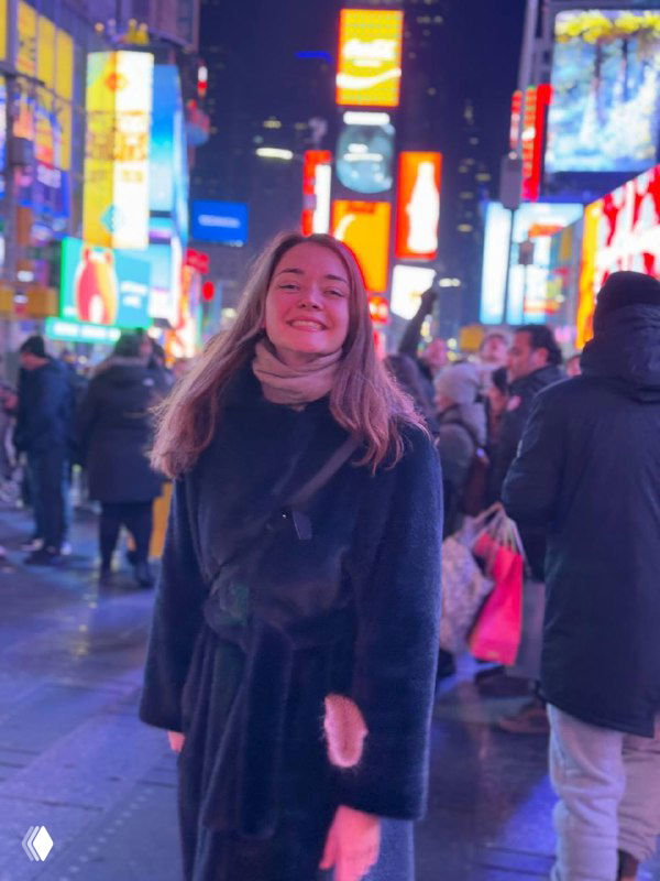 Женщина в тёмной шубе и шарфе улыбается на оживлённой ночной городской площади с яркими рекламными экранами и прохожими вокруг, атмосферное фото.