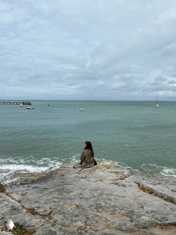 На фото женщина в пальто сидит на скалах у моря и смотрит на воду; пасмурное небо, спокойная гладь и лодки на горизонте.