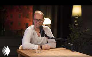 Человек в студии подкаста сидит за деревянным столом перед микрофоном; на столе стакан воды, тёплая интерьерная подсветка, кадр в спокойной обстановке.