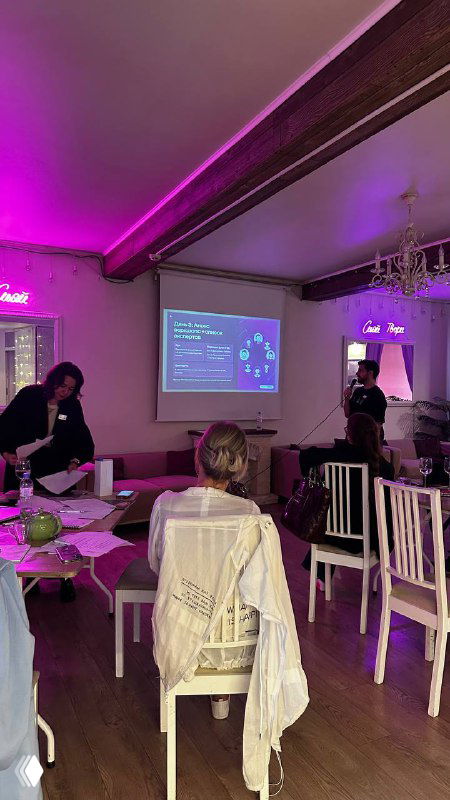 Презентация на бизнес‑игре: ведущий у экрана с слайдами, участники сидят за столами и обсуждают решения, заметна деловая атмосфера мероприятия.