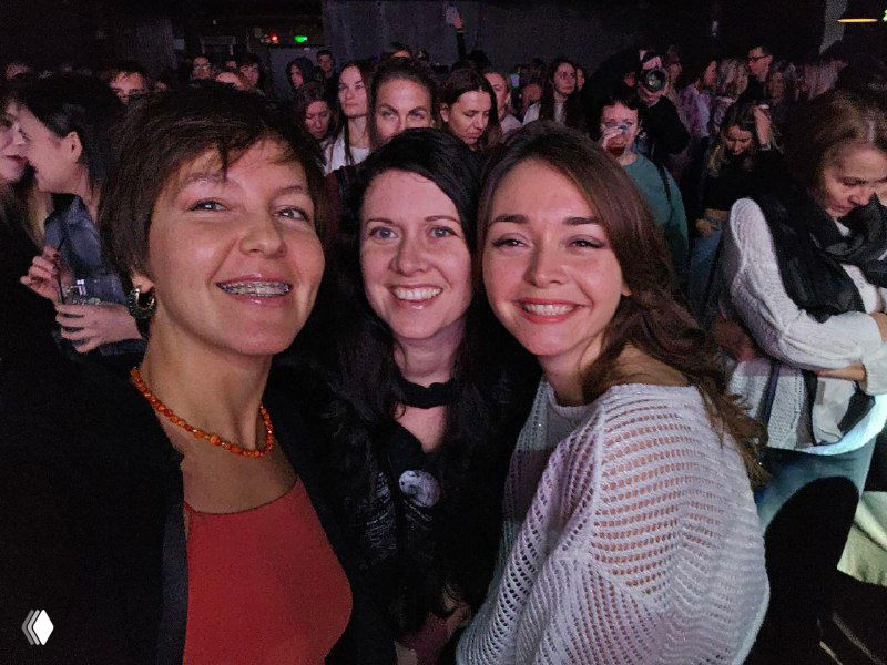 Три женщины в аудитории мероприятия улыбаются в камеру; живая офлайн‑сцена с плотной публикой на заднем плане, вечерняя теплая атмосфера общения.