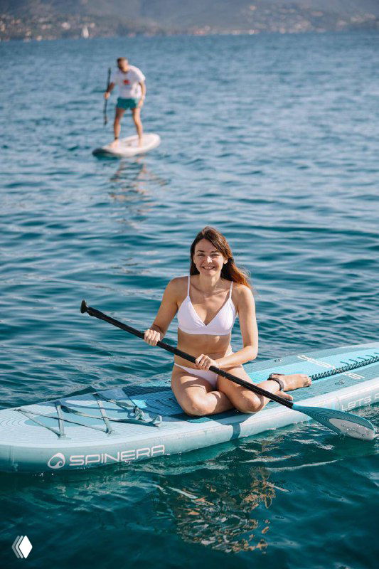 Девушка сидит на SUP-доске с веслом в руках на спокойной поверхности моря, позади виднеется городской берег и другие отдыхающие на воде.