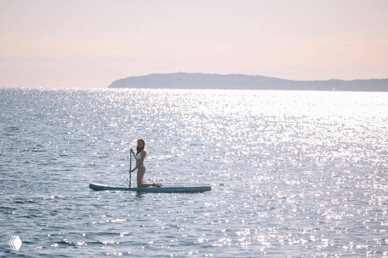 Одинокая фигура на SUP-доске в лучах солнца, море в солнечных бликах и далёкий силуэт острова; минималистичный морской пейзаж.