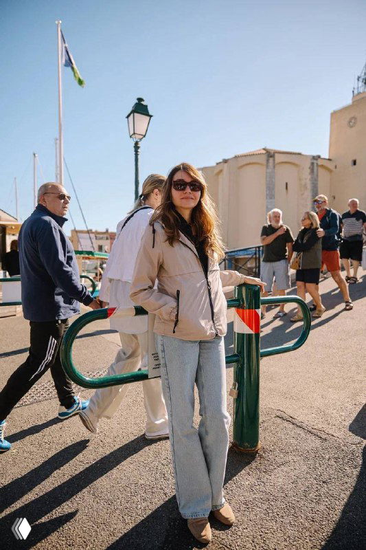 Автор идёт по набережной в лёгкой куртке и джинсах; вокруг прохожие, уличные фонари и средиземноморская архитектура городка.