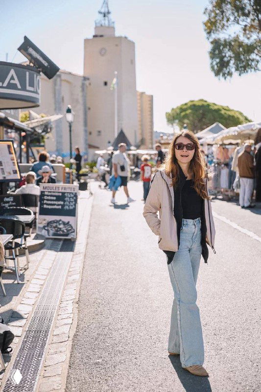 Девушка улыбается на фоне уличного рынка и кафе; солнечный день, оживлённая улица и атмосферные лавки в туристическом районе.