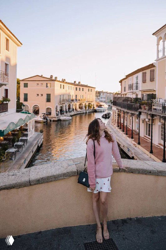 Женщина стоит на небольшом мостике над каналом, по сторонам магазинчики и дома; мягкий дневной свет и спокойная туристическая сцена.