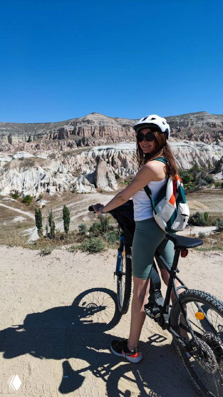 Женщина в шлеме и с рюкзаком стоит рядом с горным велосипедом на фоне скалистого пейзажа; яркий солнечный день, кадр путешествия и активности.