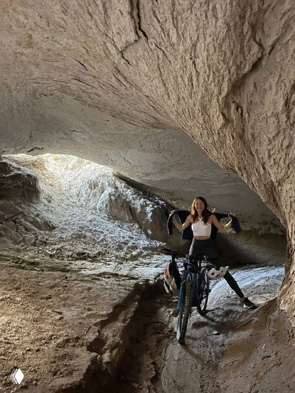 Человек с велосипедом внутри природной пещеры или грота; тонкий свет проникает в пространство, ощущение исследовательского похода и небольшого приключения.