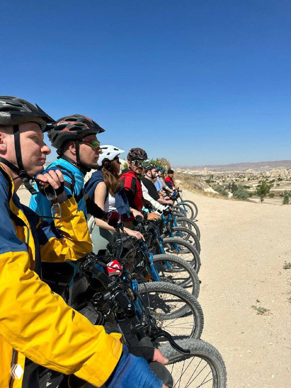 Ряд велосипедистов в защитных шлемах выстроены на горной тропе, вид на группу участников тура и просторный ландшафт на заднем плане; командный велопоход.