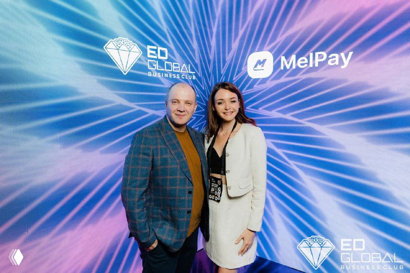 Два человека на фоне яркого баннера мероприятия с логотипами ED Global и MelPay; они в деловой одежде, позируют и улыбаются на бизнес-ивенте.
