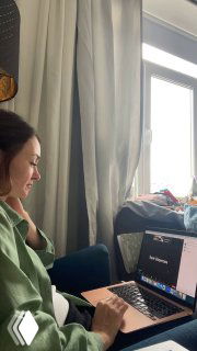 Женщина сидит за ноутбуком в домашнем рабочем кабинете; на экране видна презентация или аналитика проекта, рядом занавески и рабочие вещи.