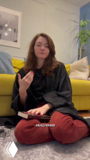 Женщина сидит на полу у жёлтого дивана с книгой на коленях задумчиво жестикулирует — визуал к теме мышления запуска процессов.