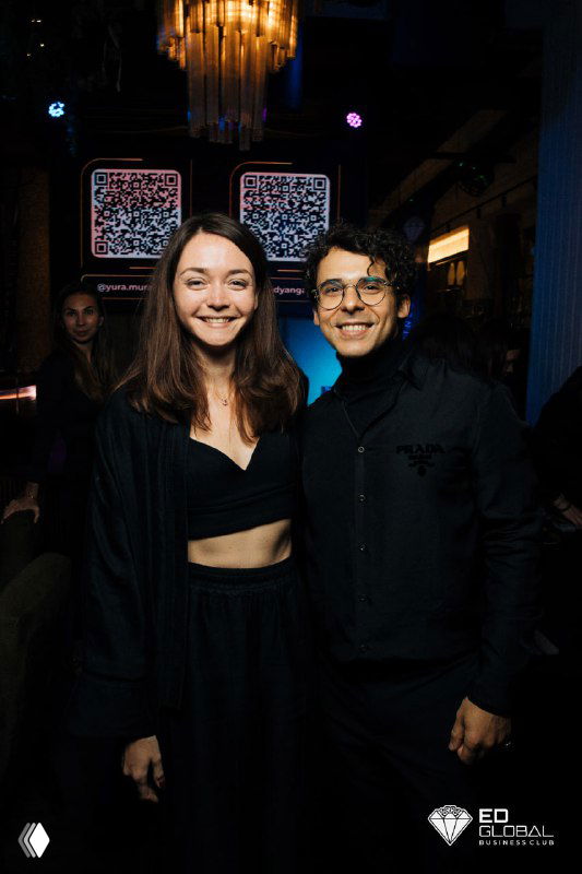 Двое людей в вечерней одежде улыбаются и позируют в тёмном интерьере мероприятия с подсветкой и декоративными панелями на заднем плане