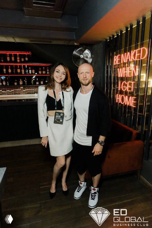 Две стоящие рядом фигуры в интерьере бара/клуба на фоне неоновой вывески и декоративного логотипа ED Global; фотография без идентификации лиц, атмосферный кадр с элементами брендинга.