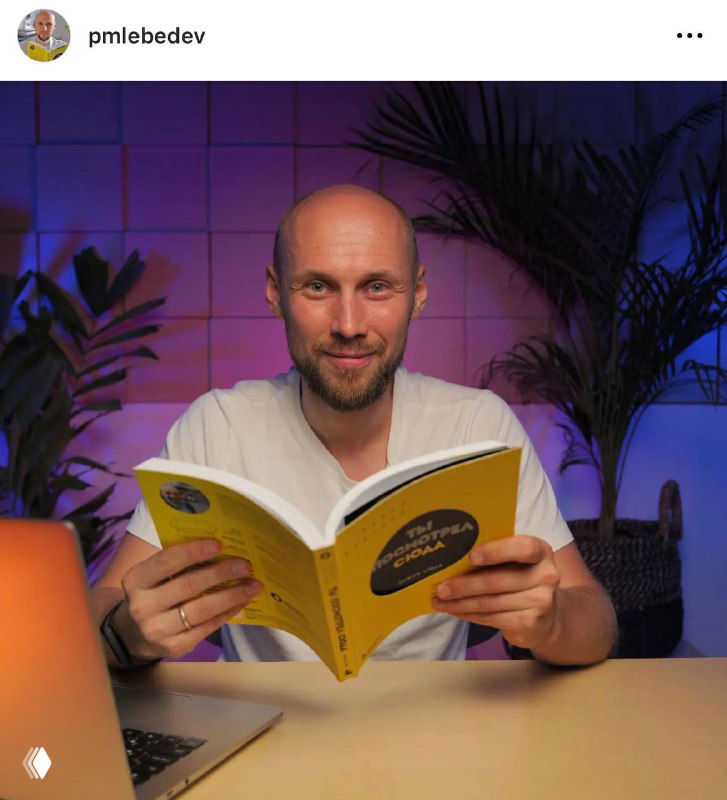 Павел Лебедев сидит за столом и улыбается, держит жёлтую книгу на фоне студийной подсветки и комнатных растений; ноутбук справа