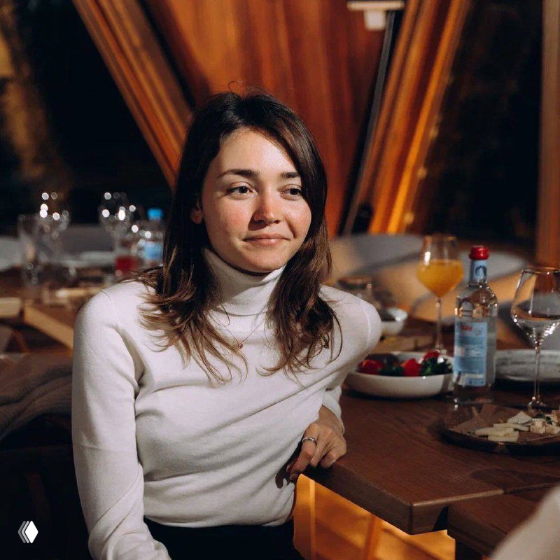 Женщина в светлом свитере сидит за обеденным столом в ресторане; на столе бокалы, бутылка воды и закуски, тёплый свет интерьера.