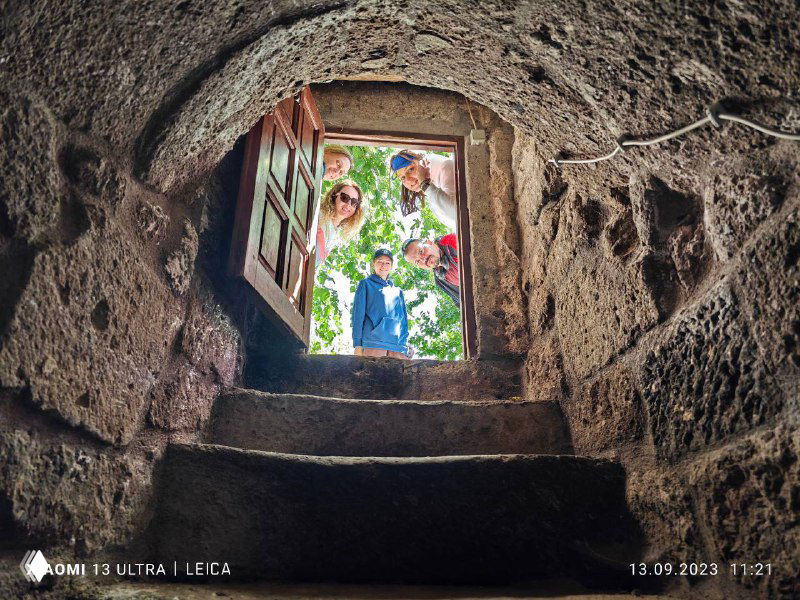 Вид снизу из каменного колодца вверх: несколько людей выглядывают в отверстие, солнечный фон и зелёная листва вокруг