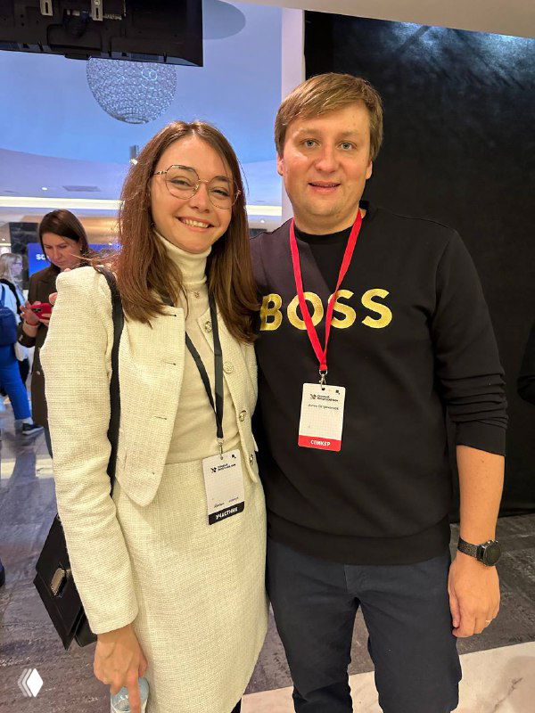 Два человека на конференции с бейджами: женщина в светлом костюме и мужчина в чёрном свитере с надписью BOSS, на заднем плане деловой зал и люди.