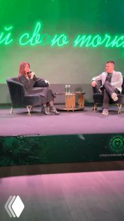 Два человека на сцене во время деловой беседы: ведущая и спикер сидят в креслах на фоне зелёного баннера, обсуждение бизнеса и разборы.