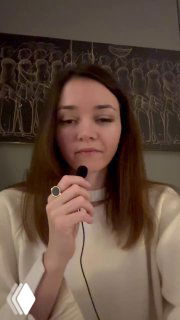 Женщина-ведущая в портретном плане держит петличный микрофон и говорит в камеру на фоне панно с контурными силуэтами людей.